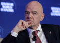 FIFA prezidentas sveikinasi su minia futbolo stadione, aplink žurnalistai su kameromis